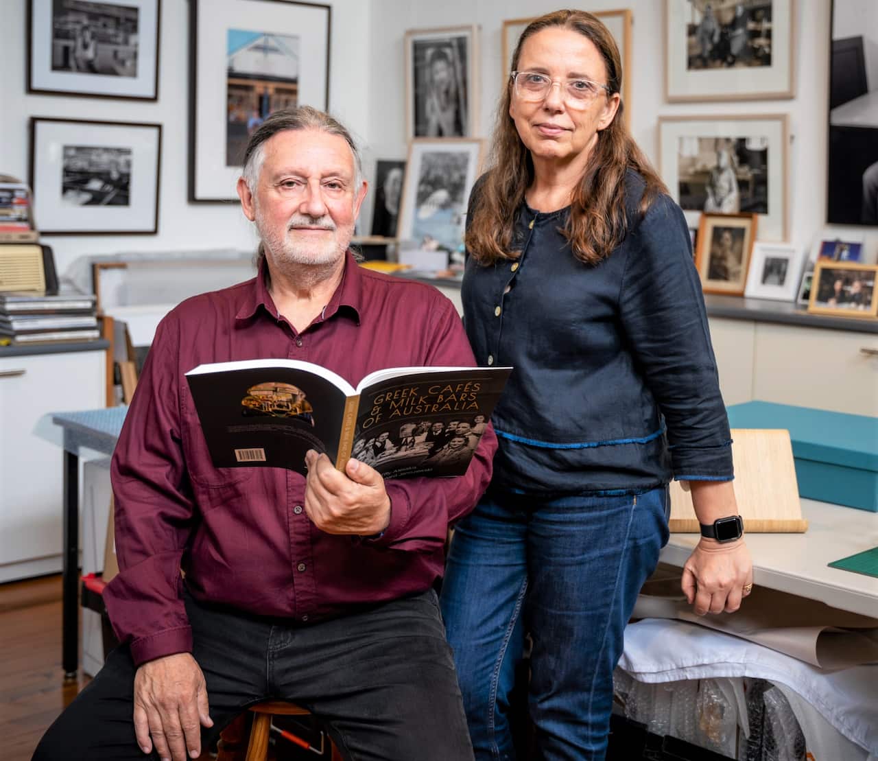 A man in a burgundy shirt sits on a stool holding an open book while a woman in a blue shirt stands beside him.