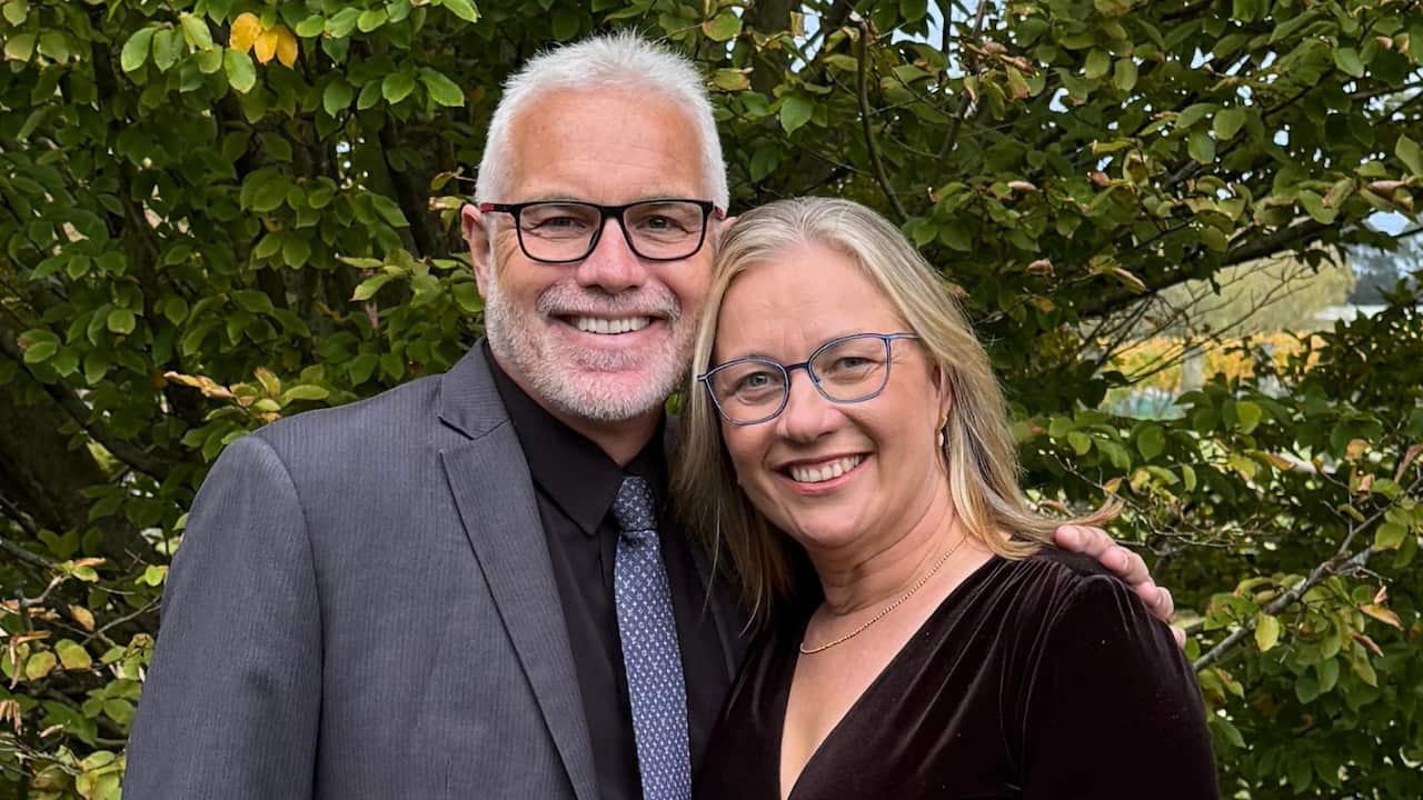 a middle aged man and woman dressed in smart casual clothing smile together in front of a tree