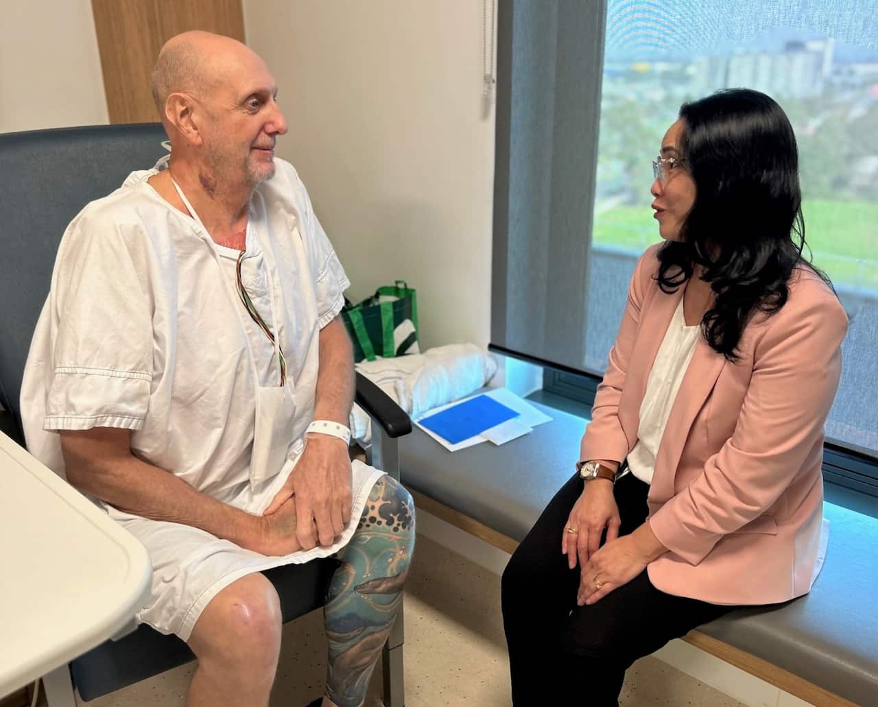 A man in a hospital gown sits in a room, talking to a woman in a pink jacket.