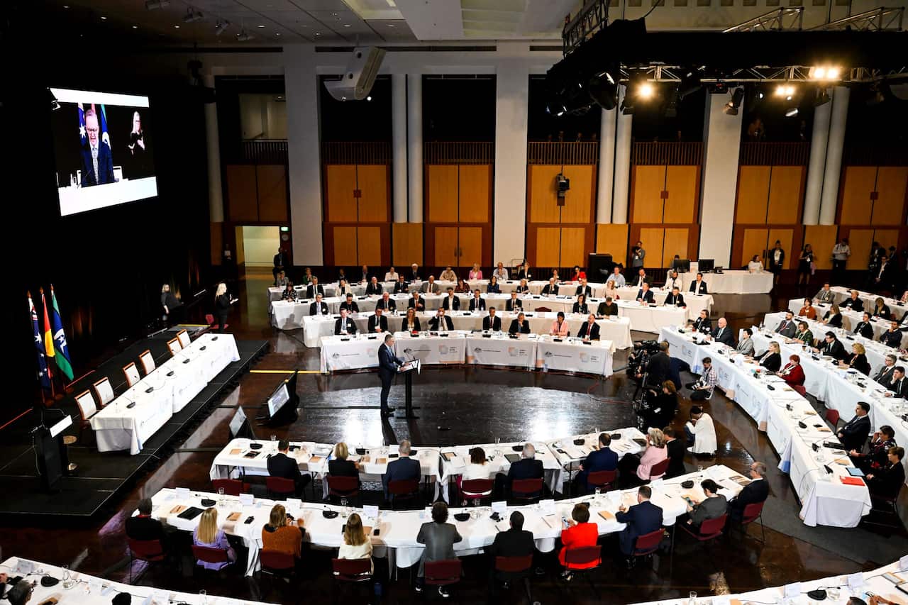 People seated at long tables. A man is speaking on a podium in the centre.