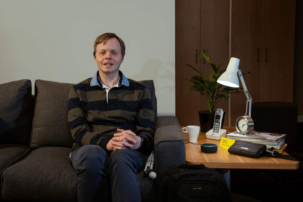 A man with a navy and green top and blue pants sits on a couch with a folded up cane next to him. On the table next to the couch, there is an Amazon Echo next to a Vision Australia sign.