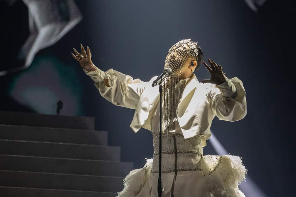 Sheldon Riley is on stage in a white ensemble, and a face covering, with his arms up. 