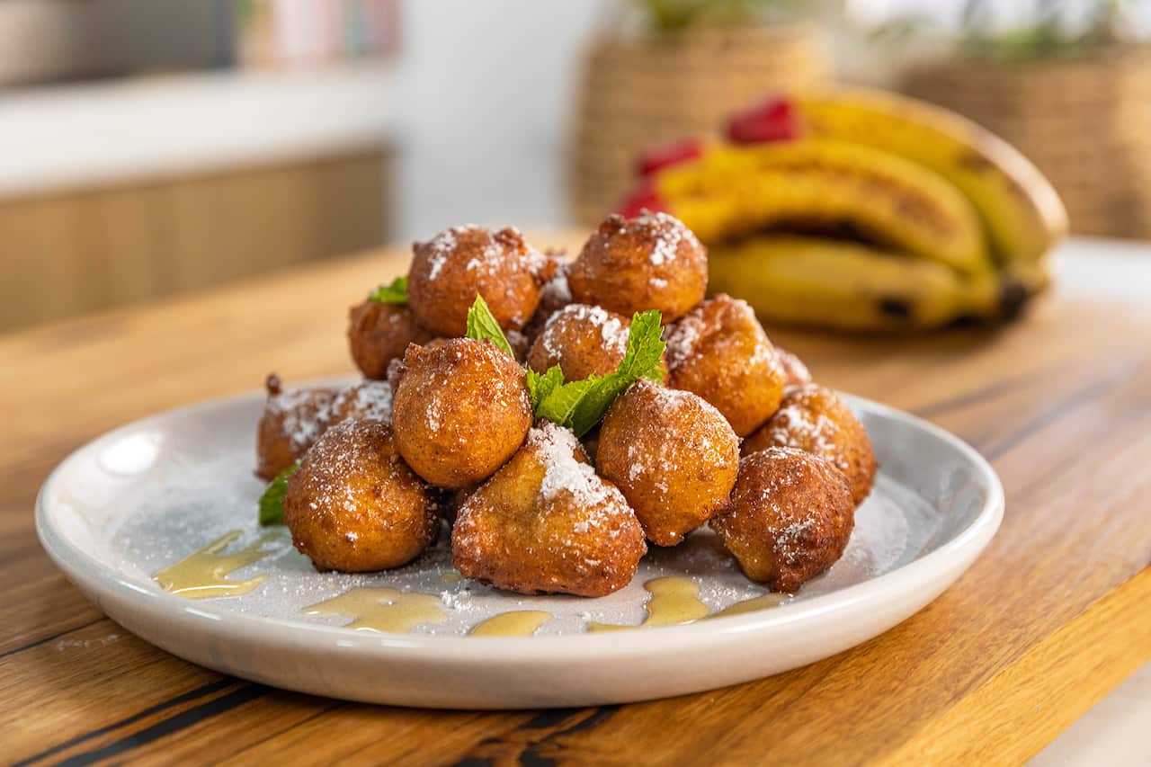 A pile of golden roundish fritters sit on a wide white plate. The fritters have been dusted with icing sugar and the plate drizzled with syrup. Bananas can be seen in the background. 