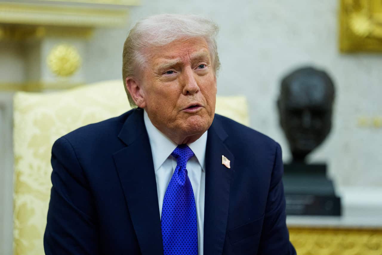 Donald Trump sitting on a golden chair, wearing a black suit, white shirt and blue tie.