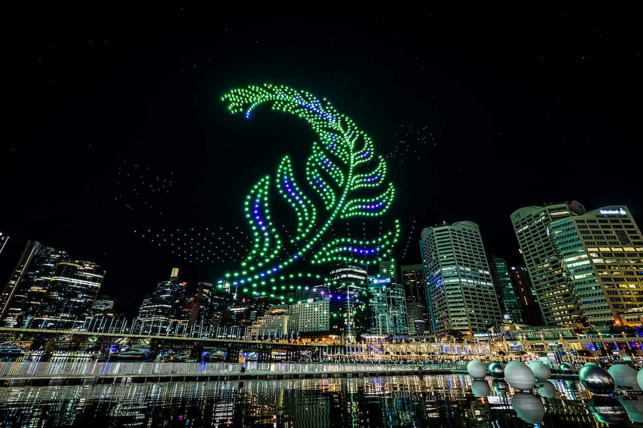 An illuminated drone light show forms a giant, glowing green fern frond over a city skyline at night, reflecting off the water below.