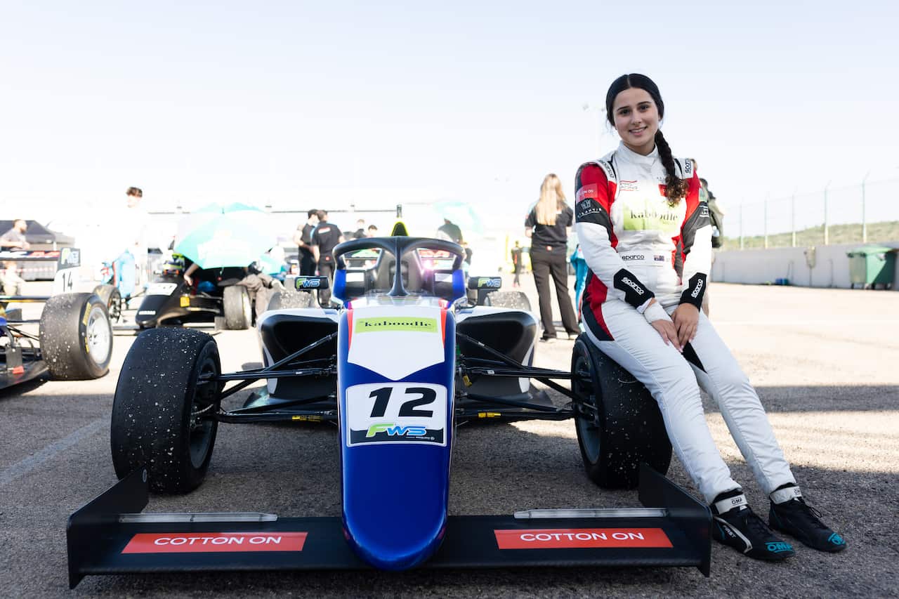 A teenage girl wearing a white racing suit sits on the front left wheel of a race car outside