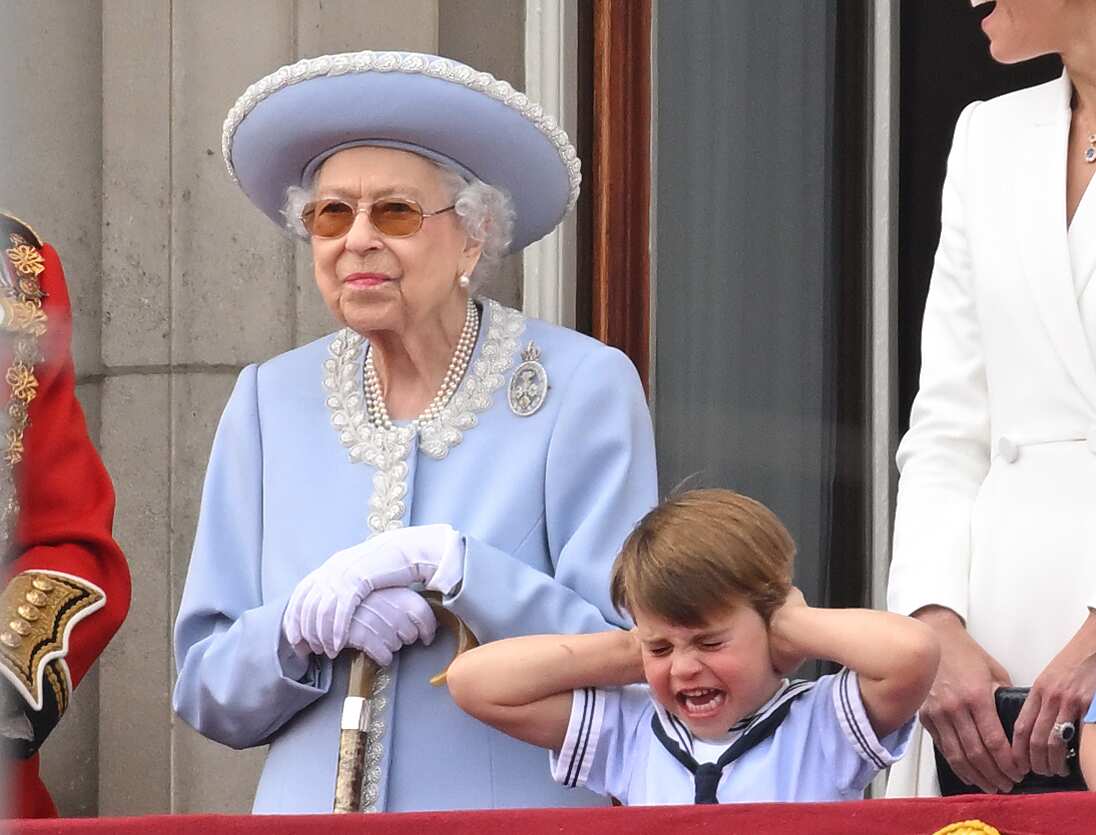 The Queen is seen standing as a boy covers his ears.