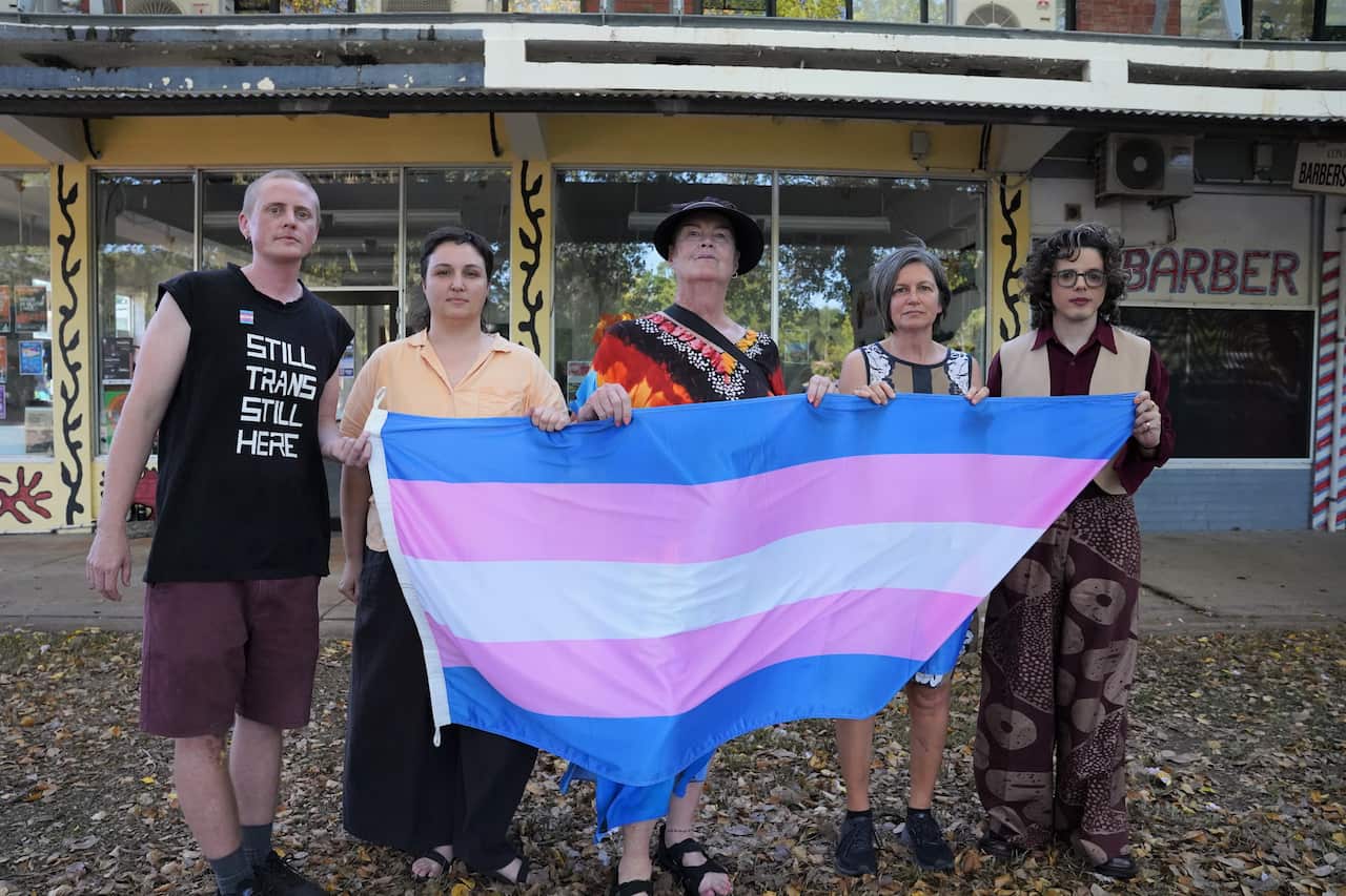 Five people standing behind and holding up a blue, pink and white transgender flag.