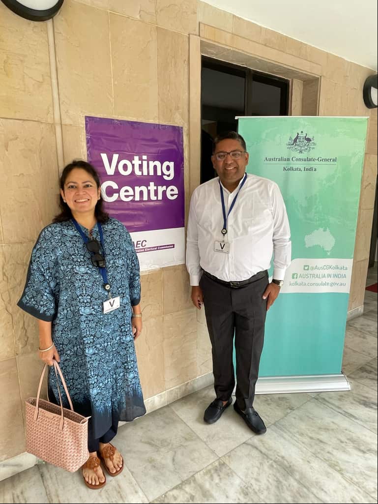 Two people stand in front of a purple voting centre sign and a teal Australian Consulate-General Kolkata, India sign.
