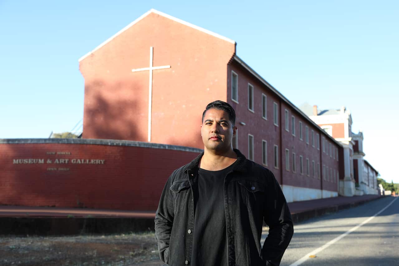 Marc Fennell, in a black shirt and jacket, stands in front of a long building. There is a cross on the end of the building, and the words 'Museum & Art Gallery' can be seen on a wall in front of the building. 