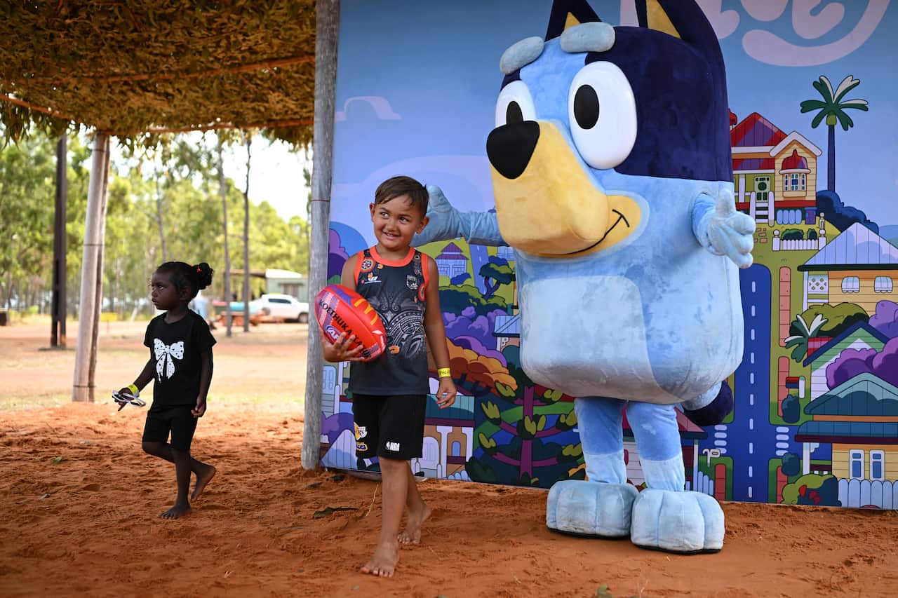 A young boy holding a football smiles as he stands outdoors next to a life-sized mascot of the cartoon character Bluey, while a young girl walks past in the background on red dirt ground.