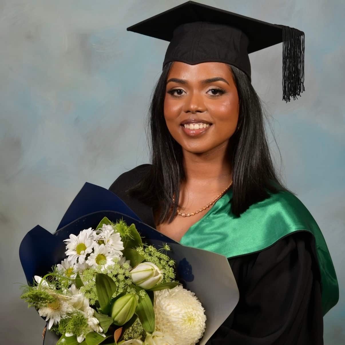 A smiling young woman in a black graduation gown, cap, and a green hood, holding a large bouquet of white flowers against a textured blue background.