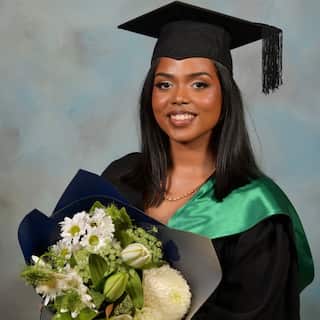 A smiling young woman in a black graduation gown, cap, and a green hood, holding a large bouquet of white flowers against a textured blue background.