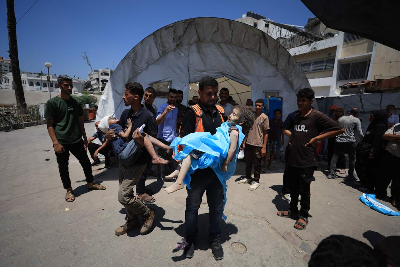 Two men carrying injured children near a large tent.
