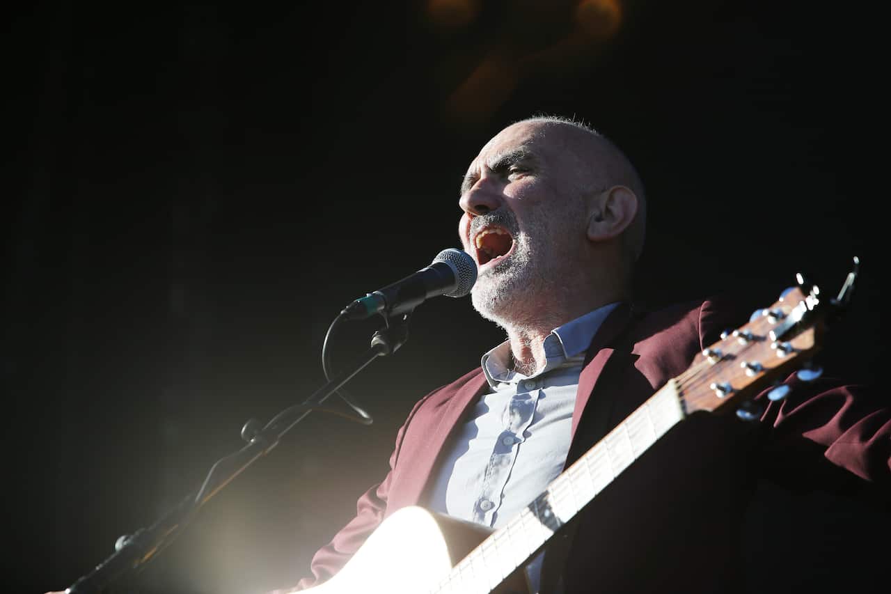 A man in a red blazer and blue shirt playing a guitar and singing into a microphone
