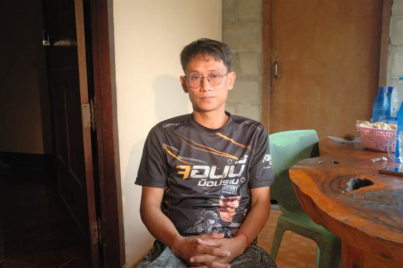 A man wearing a dark grey t-shirt with Thai writing on it. He is sitting inside next to round natural wood table.