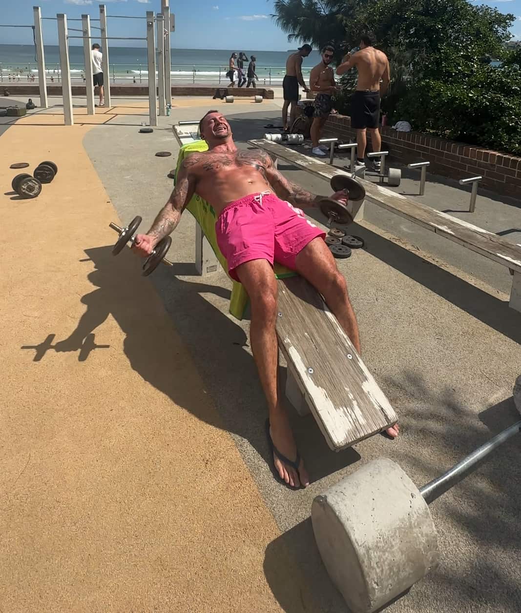 A bare-chested man in pink shorts is lifting weights on a bench in the sun