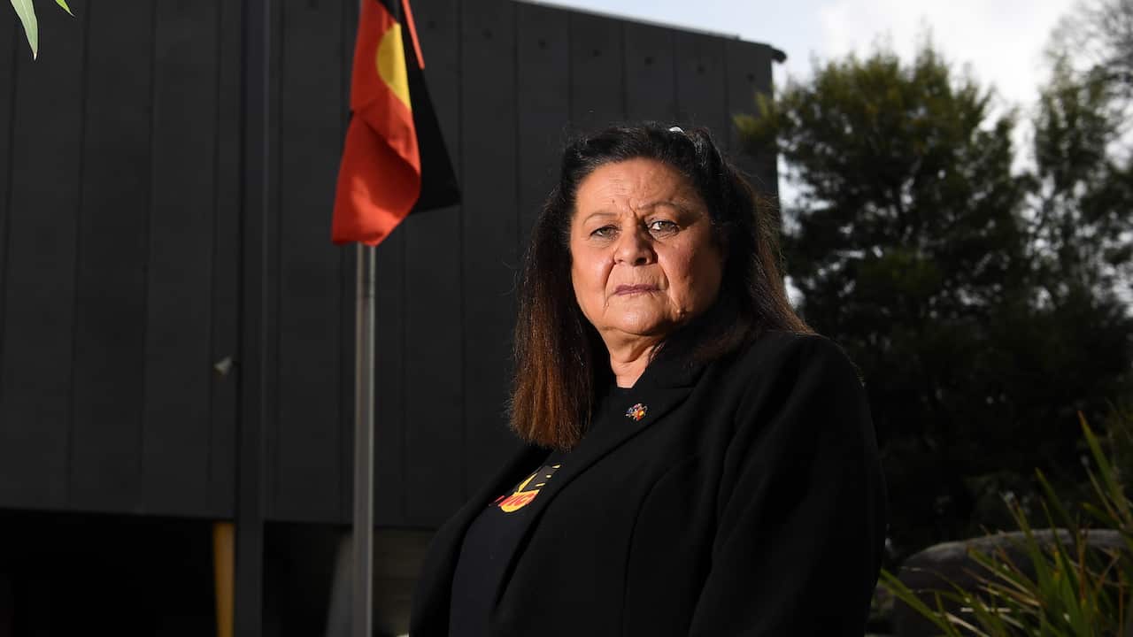 A woman wearing a blazer, standing in front of a pole with an Aboriginal flag, and a grey building behind it.
