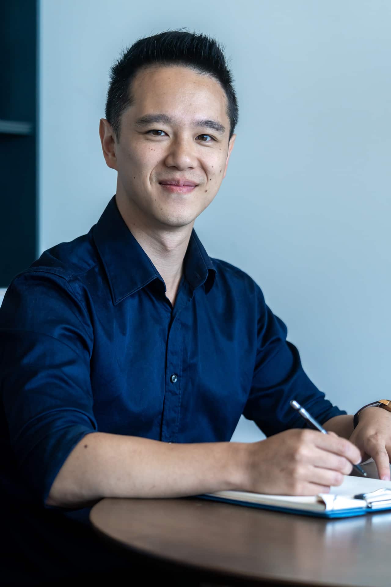 A man in a blue shirt is smiling as he writes in a notebook placed on a round wooden table in front of him.