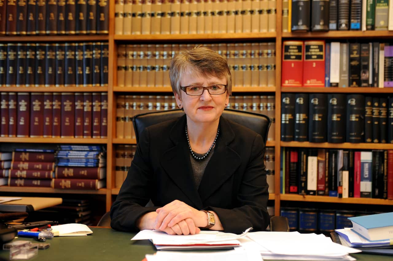 A woman with glasses and short hair, sits hands folded on a table, with law books behind her.