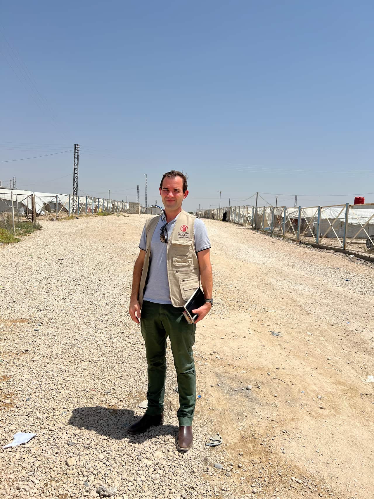 A man in a beige 'Save the Children' vest stands on a gravel pathway. To his left and right are high fences with white tents behind them. 