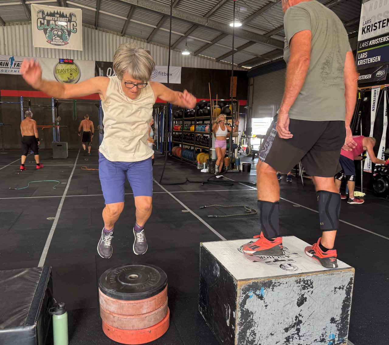 A woman in a beige singlet top jumps onto a stack of weights.
