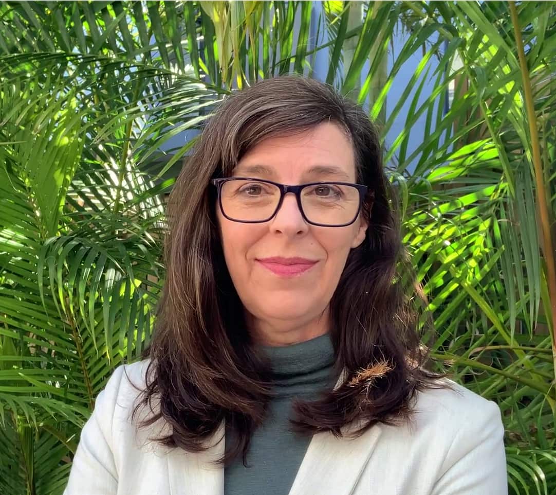 A woman with brown hair and wearing a white jacket stands in a garden setting.