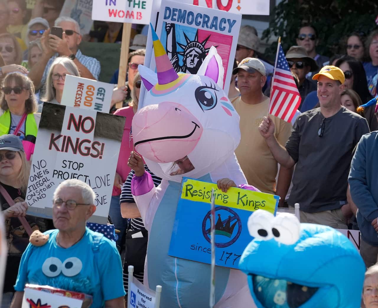 A group of people at a protest includes one person in a pink inflatable unicorn costume holding a sign that reads "Resisting Kings since 1776" and another person in the foreground wearing a blue Cookie Monster t-shirt. Other protestors hold signs reading "NO KINGS!" and "DEMOCRACY."