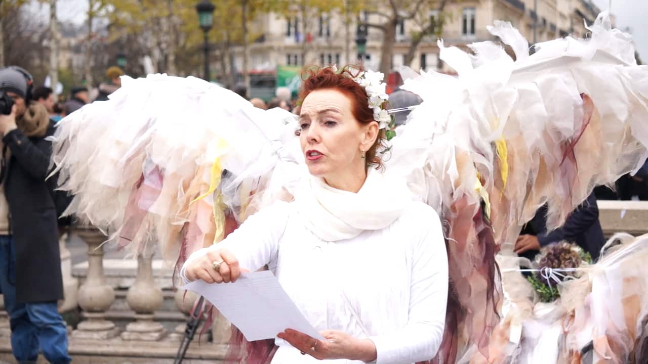 A woman with curly red hair wearing an angel costume reads from a sheet of paper held in front of her. 