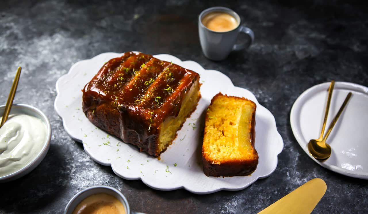 A loaf cake sits on a scallop-edged white platter. A slice has been cut off, showing a sticky top layer. The whoel cake is coated with a sticky glaze.  Cream sits in a bowl nearby. 