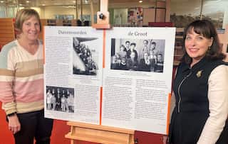 A woman weaing a striped jumper stands next to a photo board with another woman in a back vest.