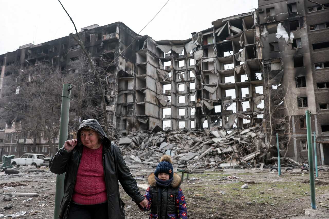 A woman holds a child's hand as they walk by a destroyed building.