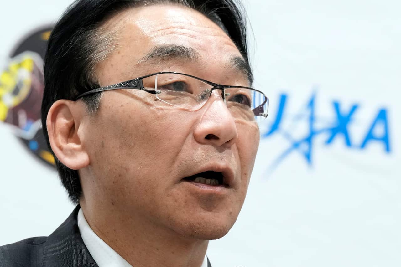 A man speaking in front of a blue JAXA logo.