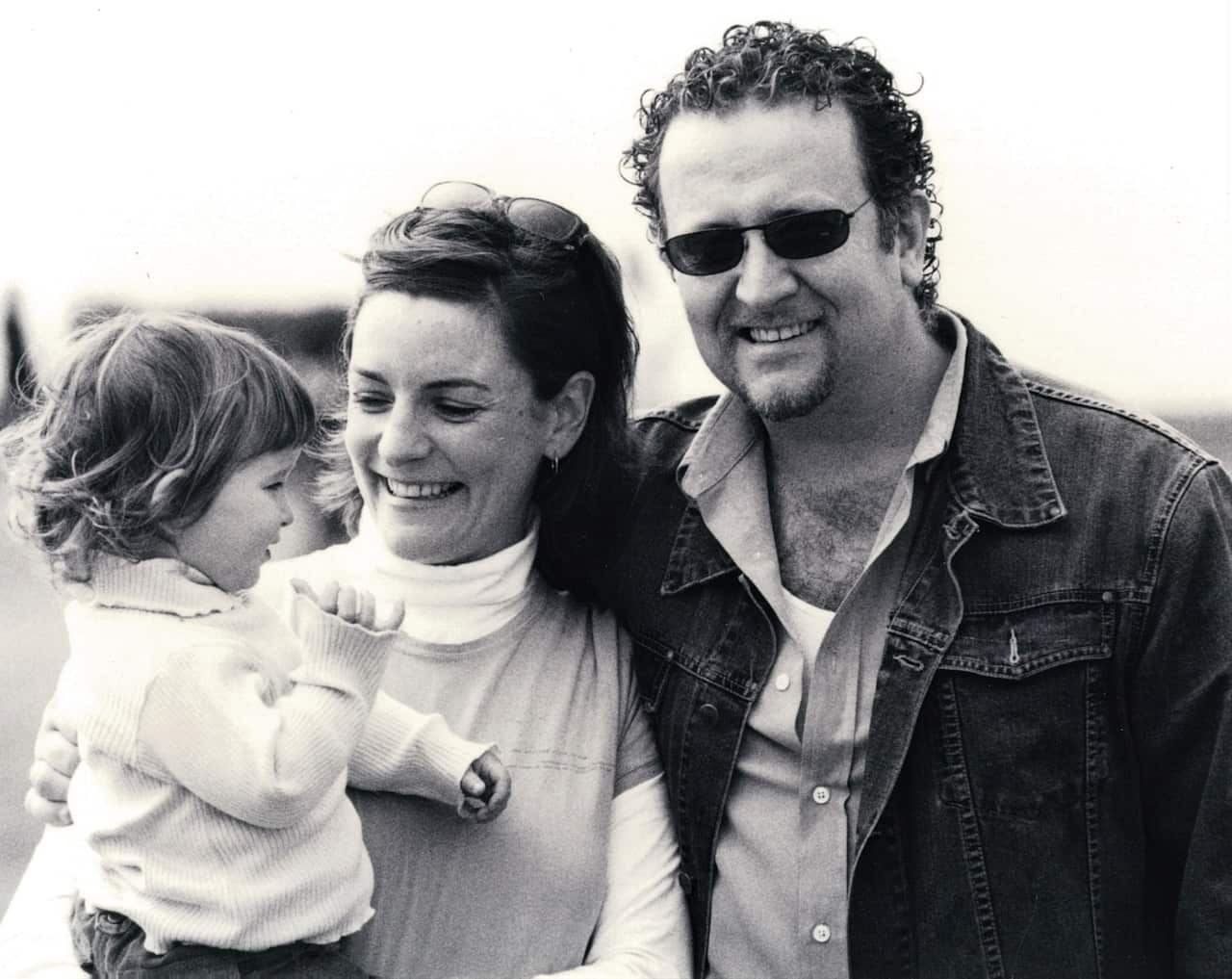 A woman looks at a small child she is holding while a man standing next to her wearing a dark denim jacket and sunnies looks at the camera.