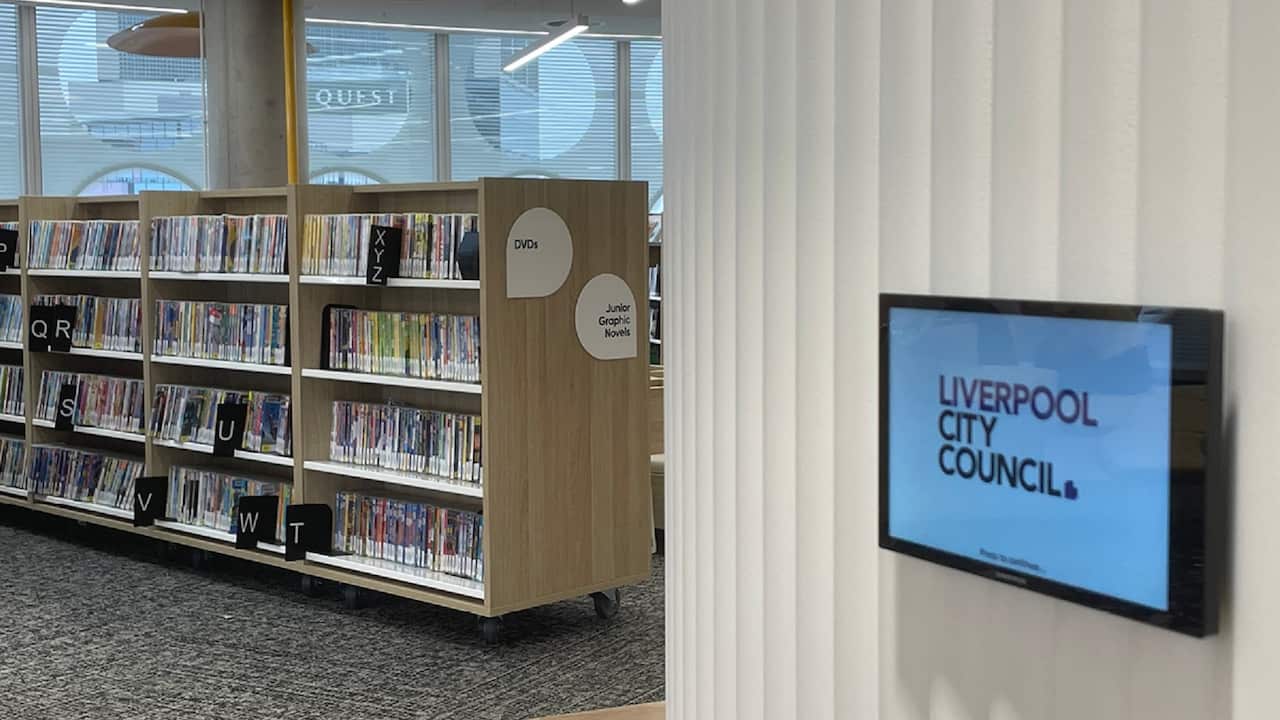 Books are arranged on shelves in a library, with a screen on the right displaying the words Liverpool City Council.