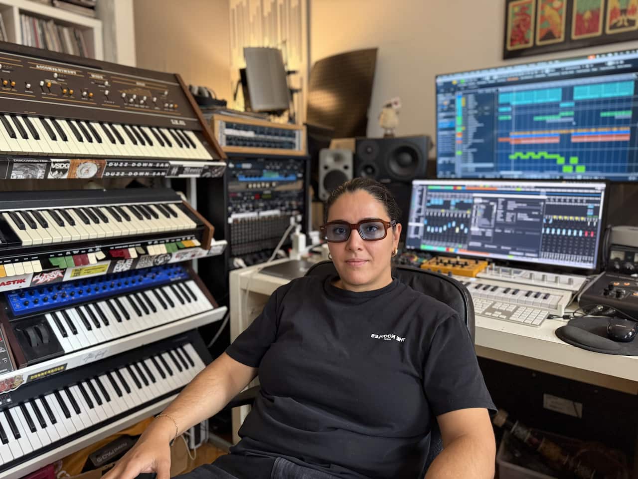 A person wearing glasses and a black R-shirt sits in a music studio surrounded by a large rack of vintage synthesisers and dual computer monitors displaying digital audio workstation software.