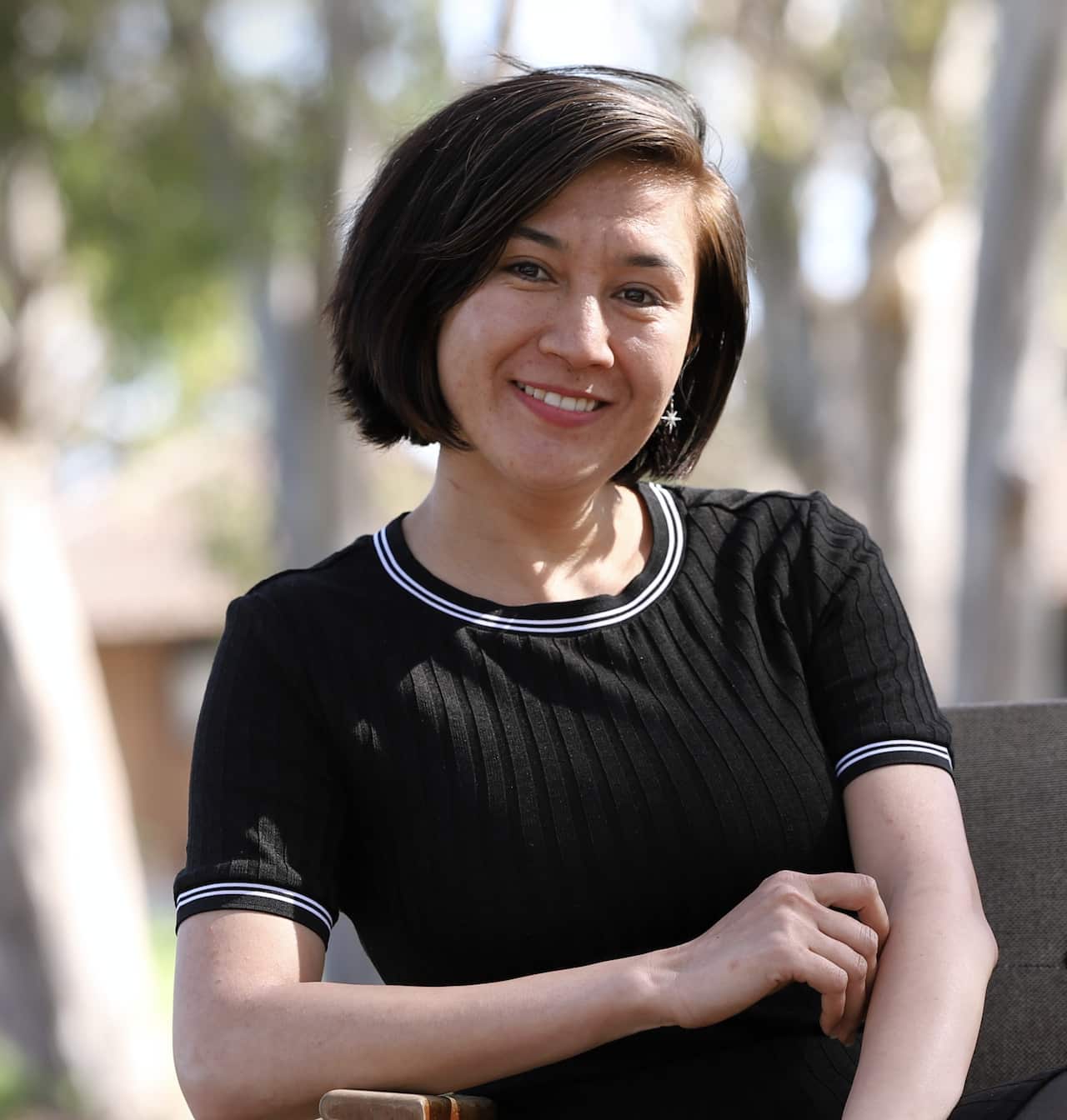 A medium shot of a smiling woman with short dark hair, wearing a ribbed black t-shirt with white striped trim, sitting outdoors with blurred trees in the background.