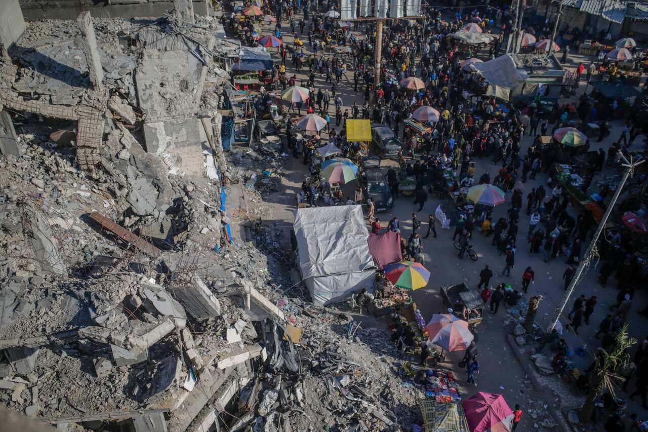 Bird's eye view shows rubble of destroyed buidings to the left of the image. On the right are crowds of people and umbrellas on the street.