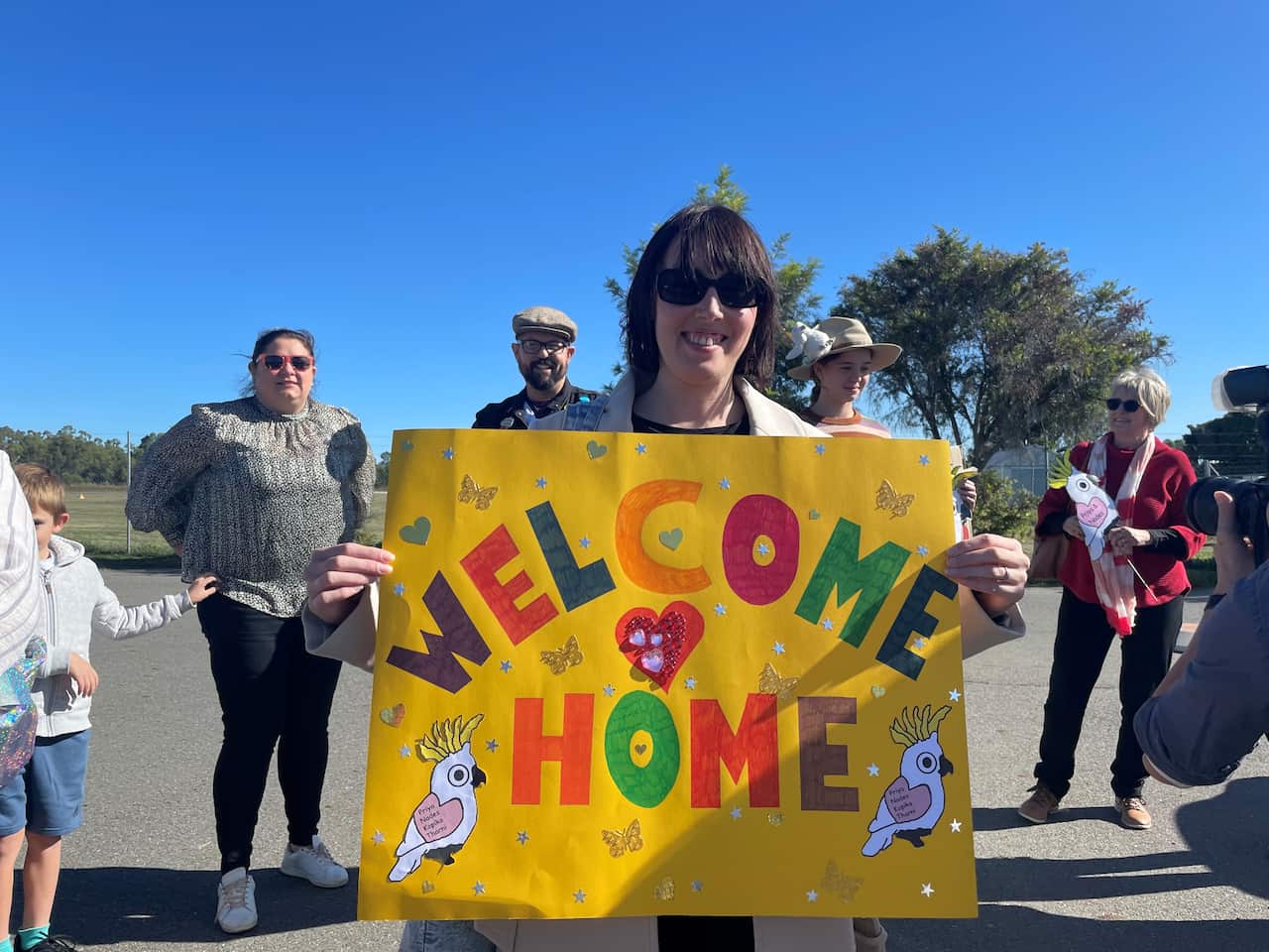 Jayne wears sunglasses, smiling carrying a yellow cardboard sign with colourful lettering: "Welcome Home".
