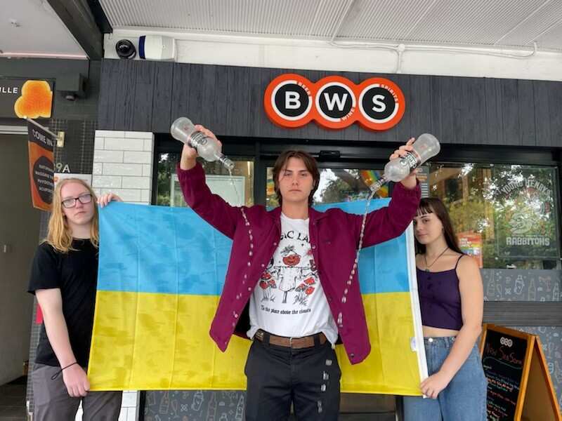 Two people hold the flag of Ukraine while another stands in front of it emptying two bottles of Russian vodka out the front of a BWS store.