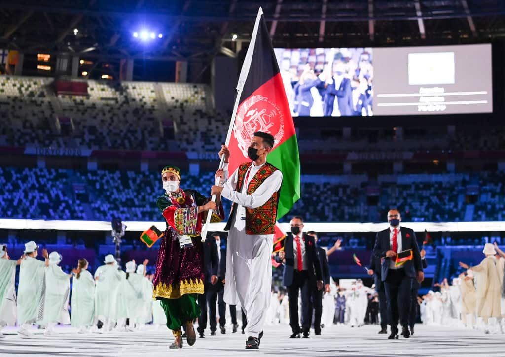 A man and a woman wearing traditional dress carry the flag of Afghanistan in a stadium