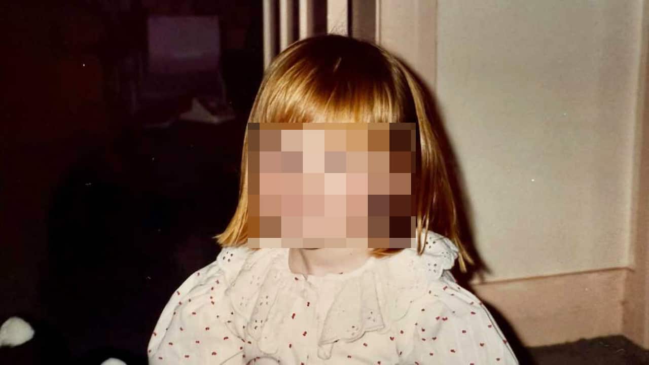 A young girl with short, red hair and a fringe stands in a house, wearing a white polka dot top with a frilly collar. Her face is pixelated.