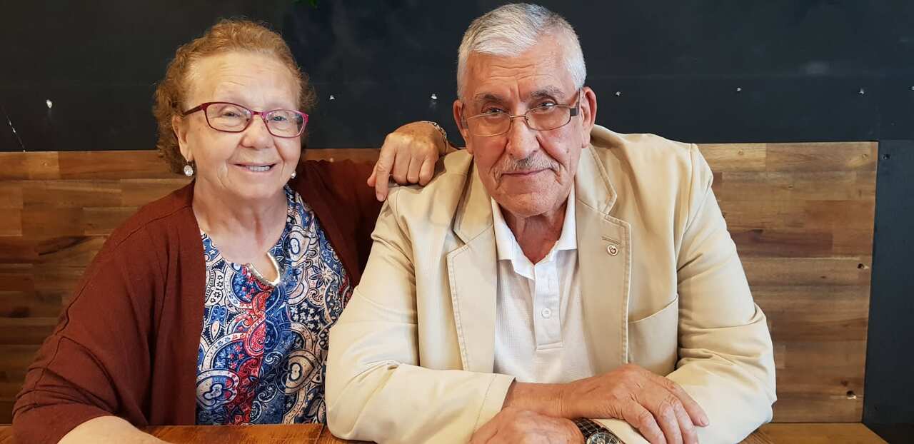 An elderly couple sit together at a wooden table and smile as they face the camera.