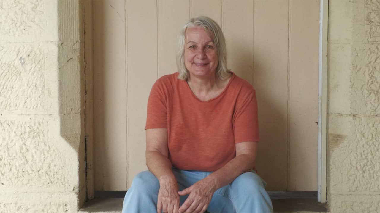an older woman in a orange tshirt and blue jeans sitting on a step outside a door