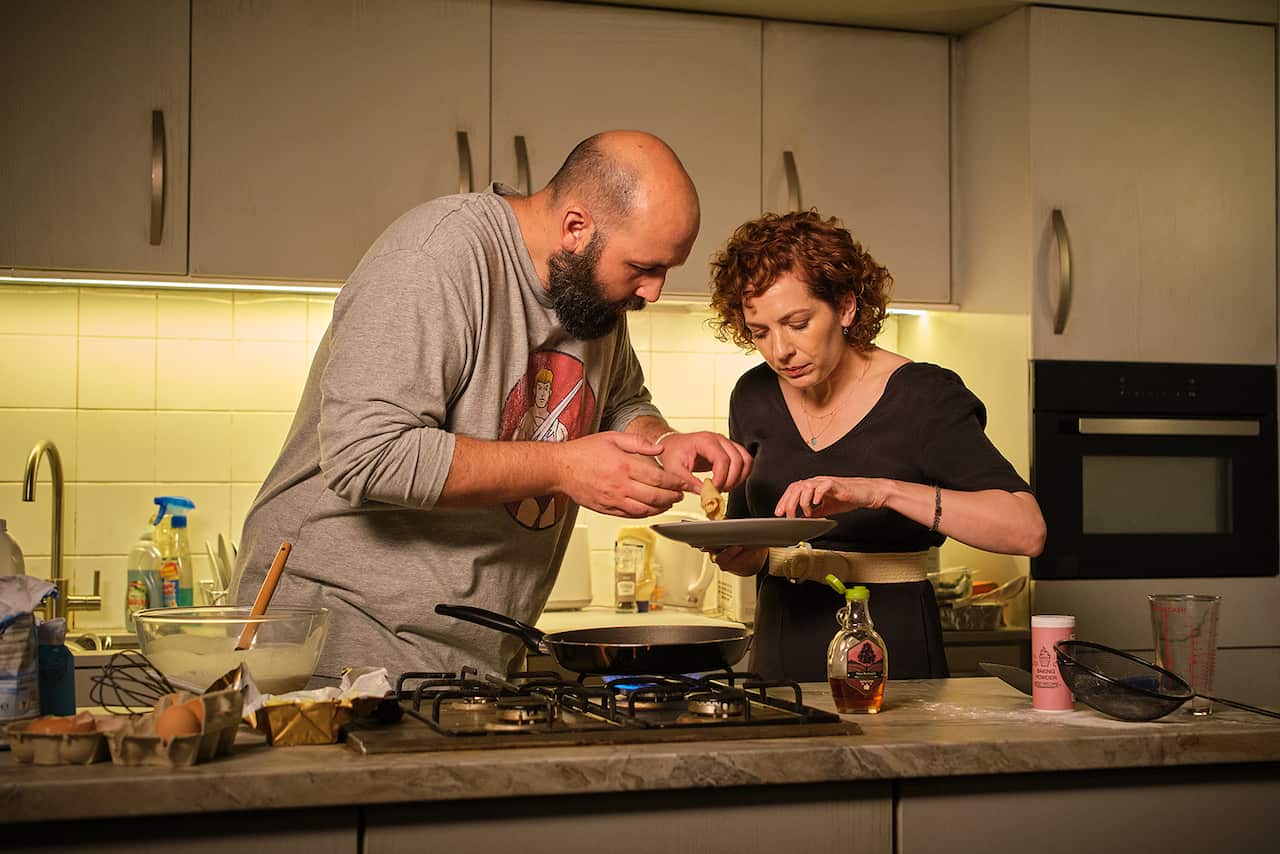 A man and a woman are in a kitchen, standing behind a cooktop in a bench. They are both leaning over a plate that she holds. 