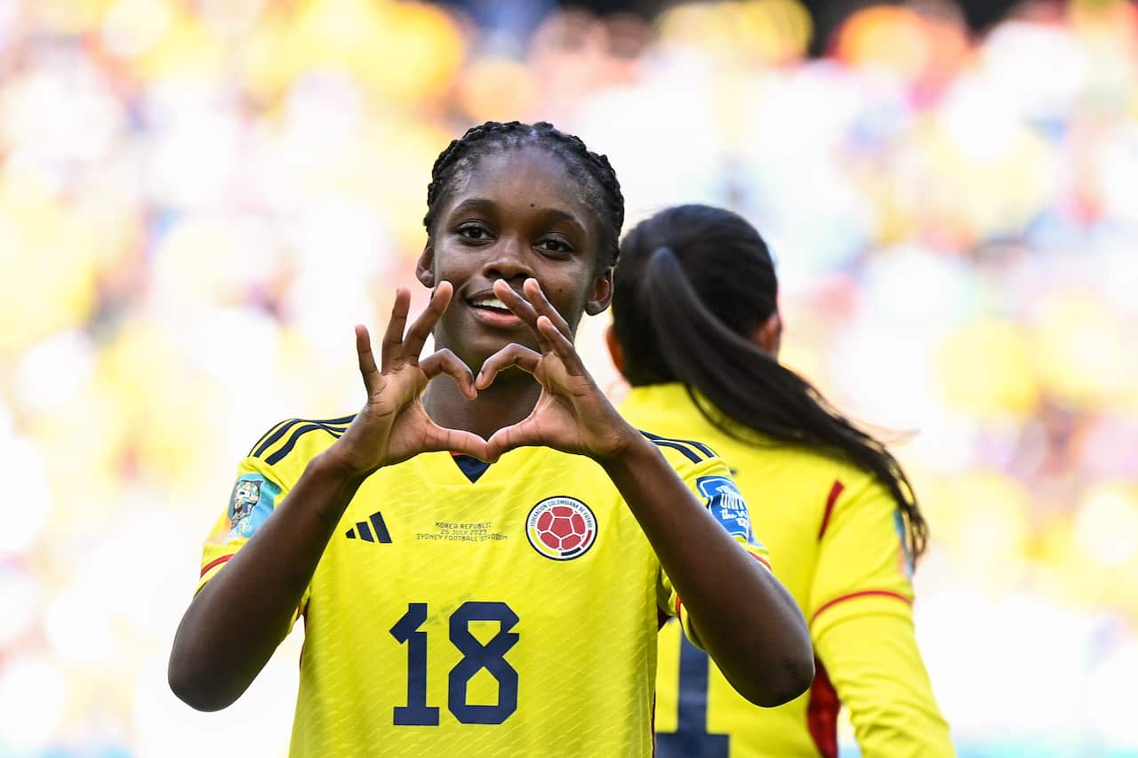 A woman in a yellow football jersey making a heart sign with her hands.