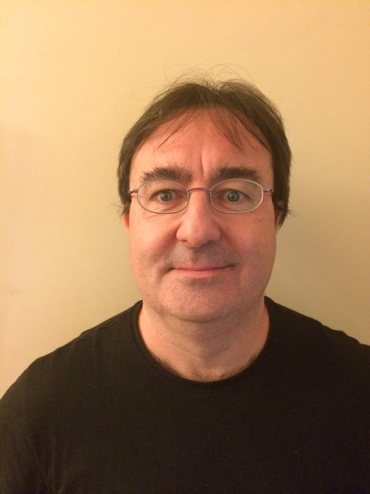 A man standing against a yellow wall. He's wearing glasses and a black T-Shirt