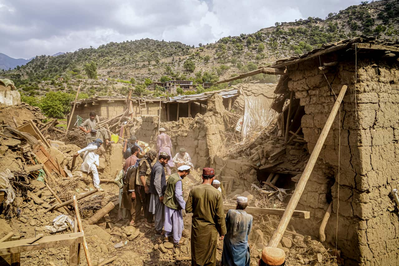 A group of people searching through rubble, with severely damaged buildings surrounding them.