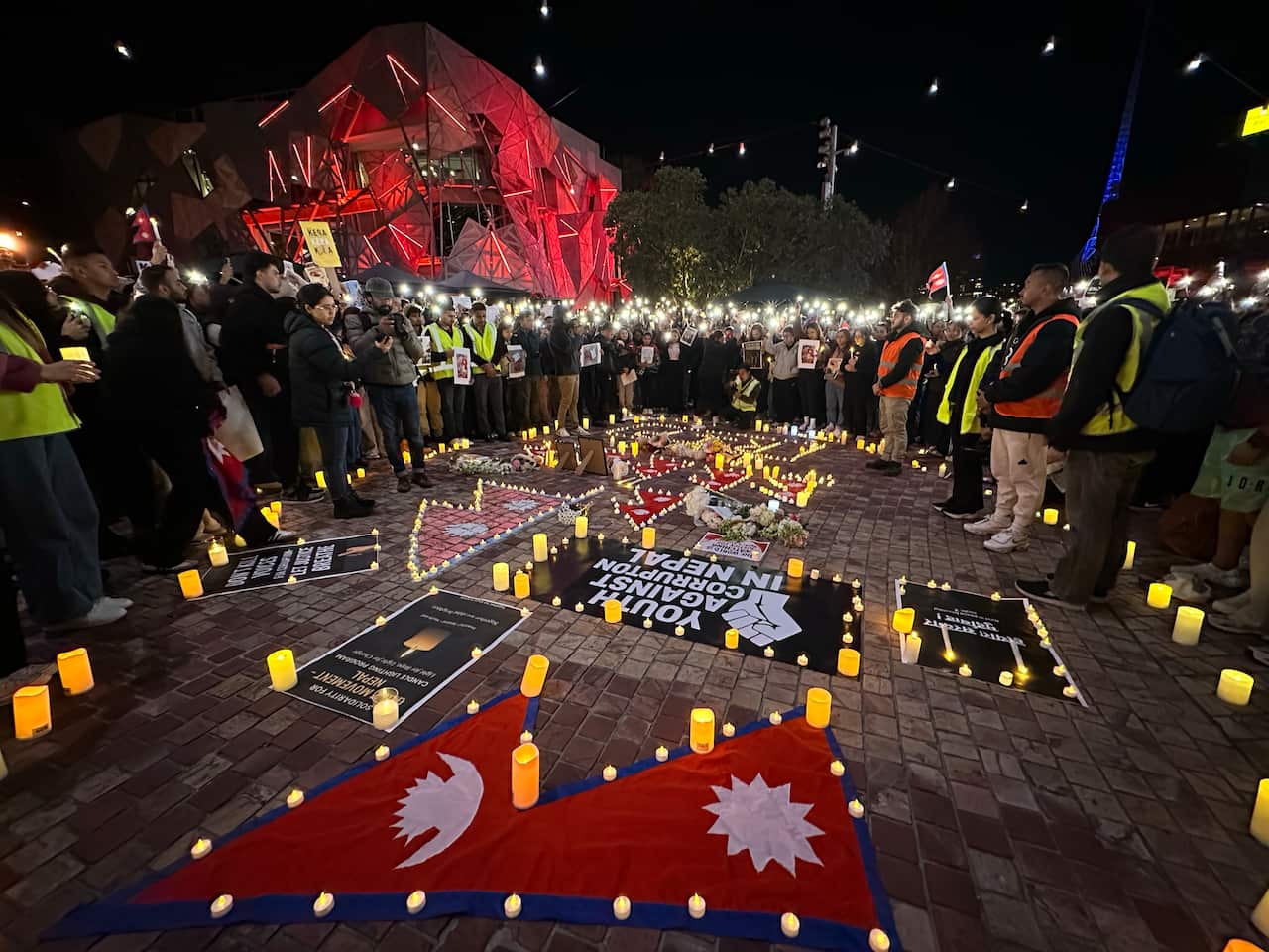 A nighttime photo shows a group of people standing around a large display of candles and signs on a cobblestone plaza. The Nepali flag is laid out on the ground, surrounded by protest signs and candles, while many people in the background hold up their phones with flashlights on.