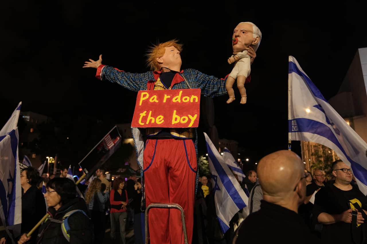 A person dressed as Donald Trump at a rally. He is wearing a sign reading 'Pardon the Boy' around his neck, while holding up a doll meant to represent Benjamin Netanyahu